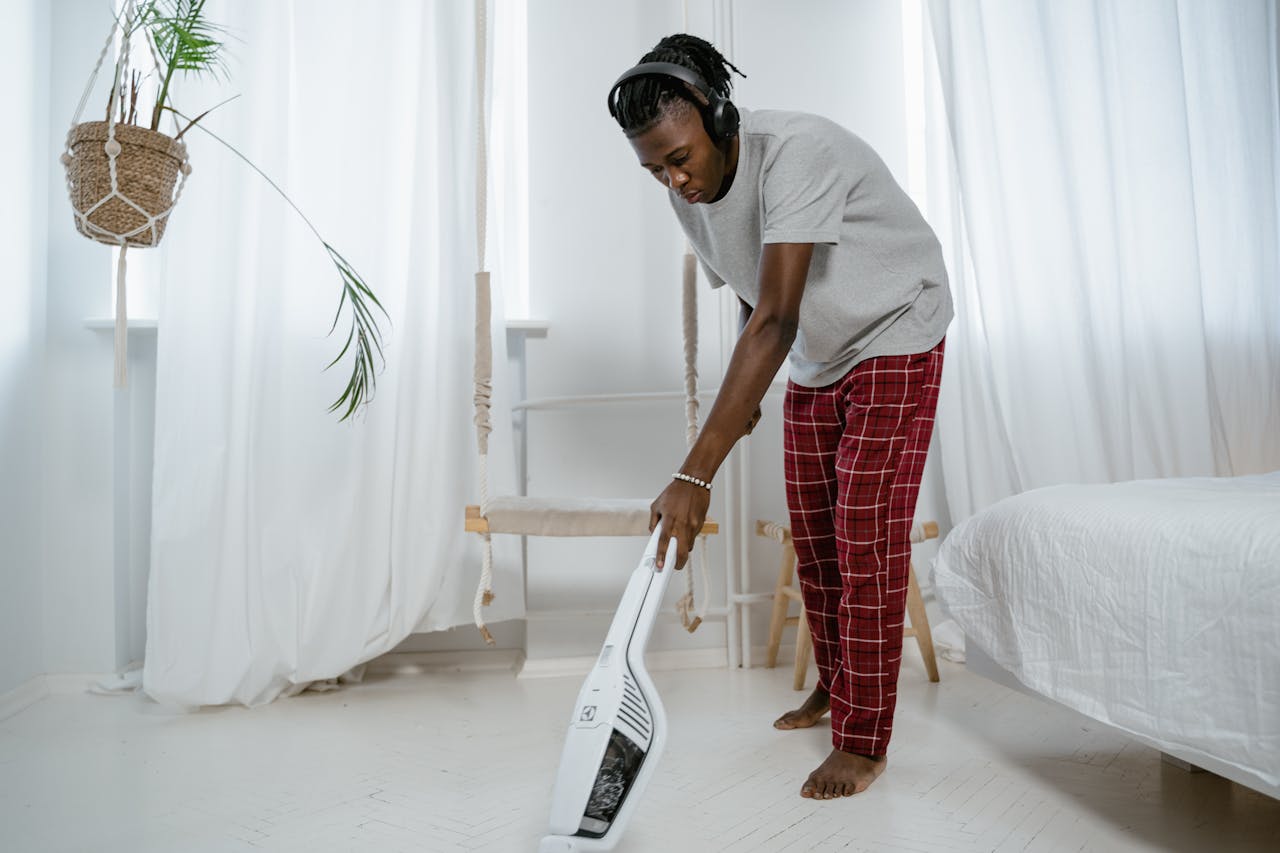 A man performs house chores, vacuuming in a stylish bedroom with natural light.